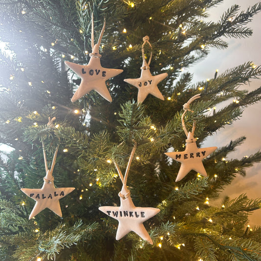 Handmade ceramic star ornaments by Betsey Carter with leather ribbon, featuring holiday words: "Merry," "Love," "Joy," "Fa La La," and "Twinkle."