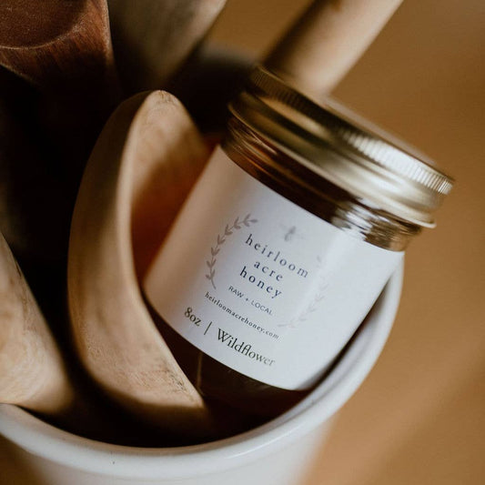 A jar of Wildflower Honey by Heirloom Acre Honey, 8 oz, raw and unfiltered with a rustic label, sitting next to a honey dipper and wildflowers.